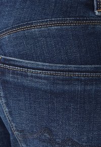 Jeans in denim blu scuro con cuciture a contrasto, dotati di una tasca posteriore e dettagli di cucitura curva subtle. La texture sembra robusta e durevole.