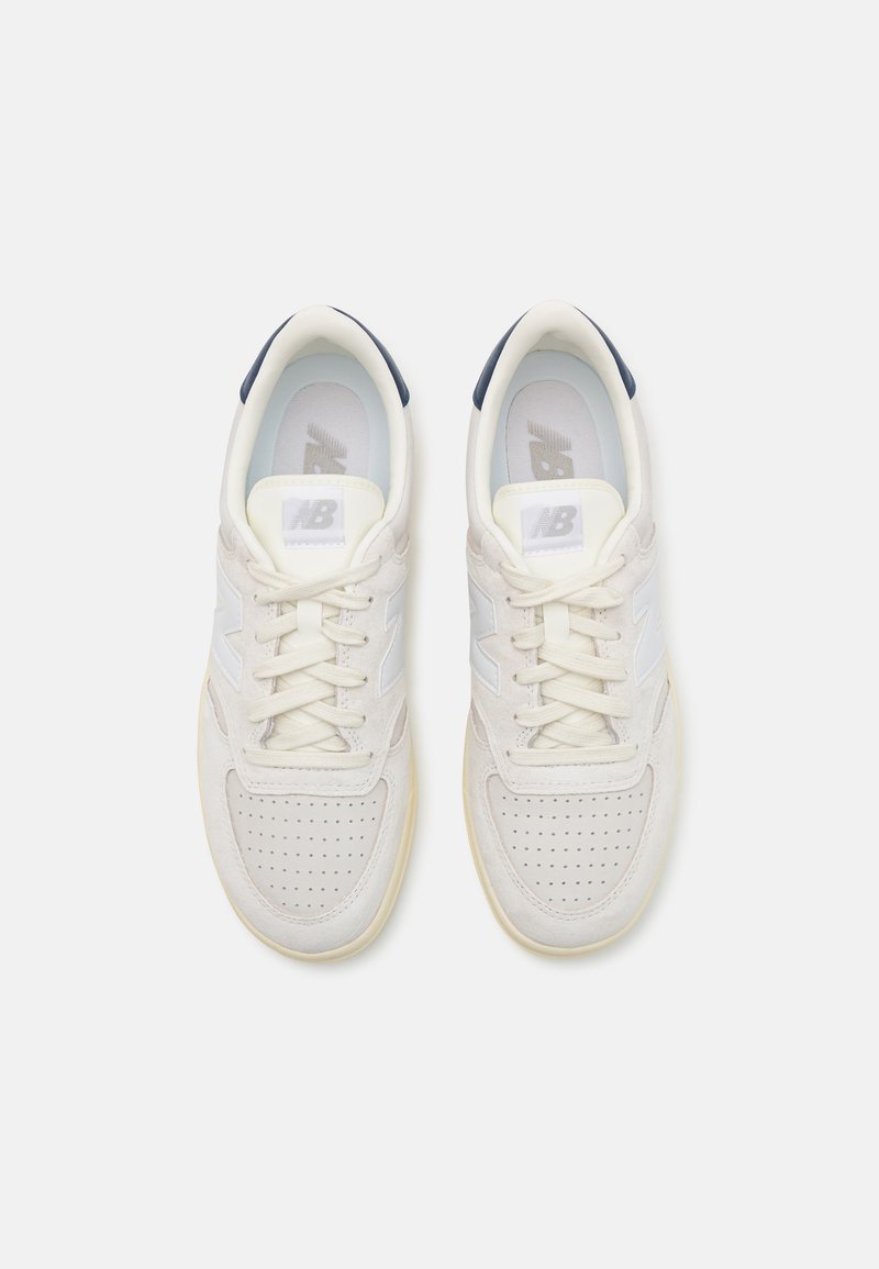 Zapatillas de ante blanco con detalles de malla, puntera perforada, cierre de cordones y acentos en el talón en color marino. Presentan el logo "NB" en gris reflectante.