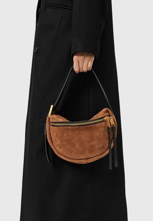 Brun ruskindshåndtaske med buet form, der har en gylden lynlås og sort læderhåndtag, båret af en person i en sort frakke.