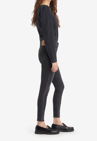 Mujer de pie con una blusa de manga larga de mezclilla negra, jeans ajustados negros con dobladillos sin acabado y mocasines negros contra un fondo blanco.
