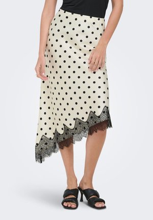 Femme portant une jupe midi crème à pois noirs avec bordure en dentelle, associée à un haut noir sans manches et des sandales à talons noires.