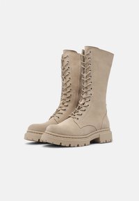 Beige suede knee-high combat boots with a lace-up front, metal eyelets, thick rubber sole, and rounded toe. Textured finish enhances grip.