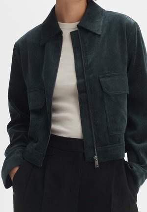 Persoon in een donkergroene ribfluwelen jack met voorzakken over een witte geribbelde shirt en zwarte high-waisted broek.