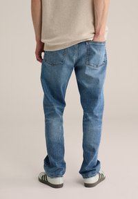 Mėlynos spalvos džinsai iš džinsinio audinio, laisvo silueto, su nugaros kišenėmis, šiek tiek nuvalytu efektu ir dėvėtu paviršiumi, derinami su šviesių spalvų batais.