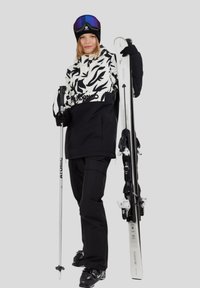 Donna in un completo da sci bianco e nero che tiene gli sci e un bastone da sci, indossando guanti neri, occhiali da sci e un berretto su uno sfondo neutro.