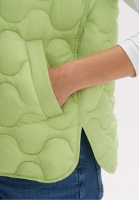 Pikowana zielona kamizelka puchowa o gładkiej fakturze, z boczną kieszenią i zaokrąglonym dołem. Materiał wydaje się lekki i ciepły.