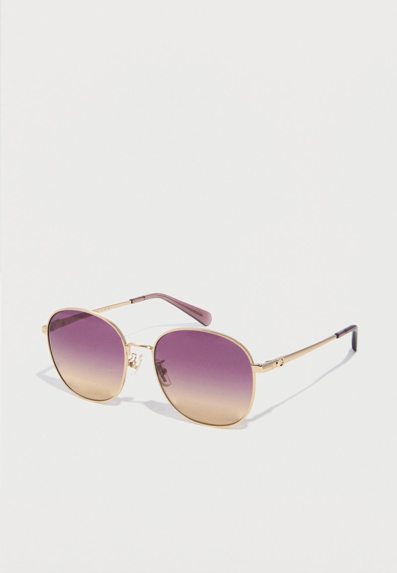 Γυαλιά ηλίου με χρυσή κορνίζα, στρογγυλοί τετράγωνοι φακοί με μοβ διαβάθμιση και λεπτά μπράτσα που ακουμπούν σε λευκή επιφάνεια.