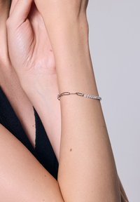 Srebrna bransoletka z wzorem łańcuchowym, ozdobiona rzędami małych, okrągłych, przezroczystych kamieni, spoczywająca na nadgarstku. Gładka faktura, delikatny wygląd.