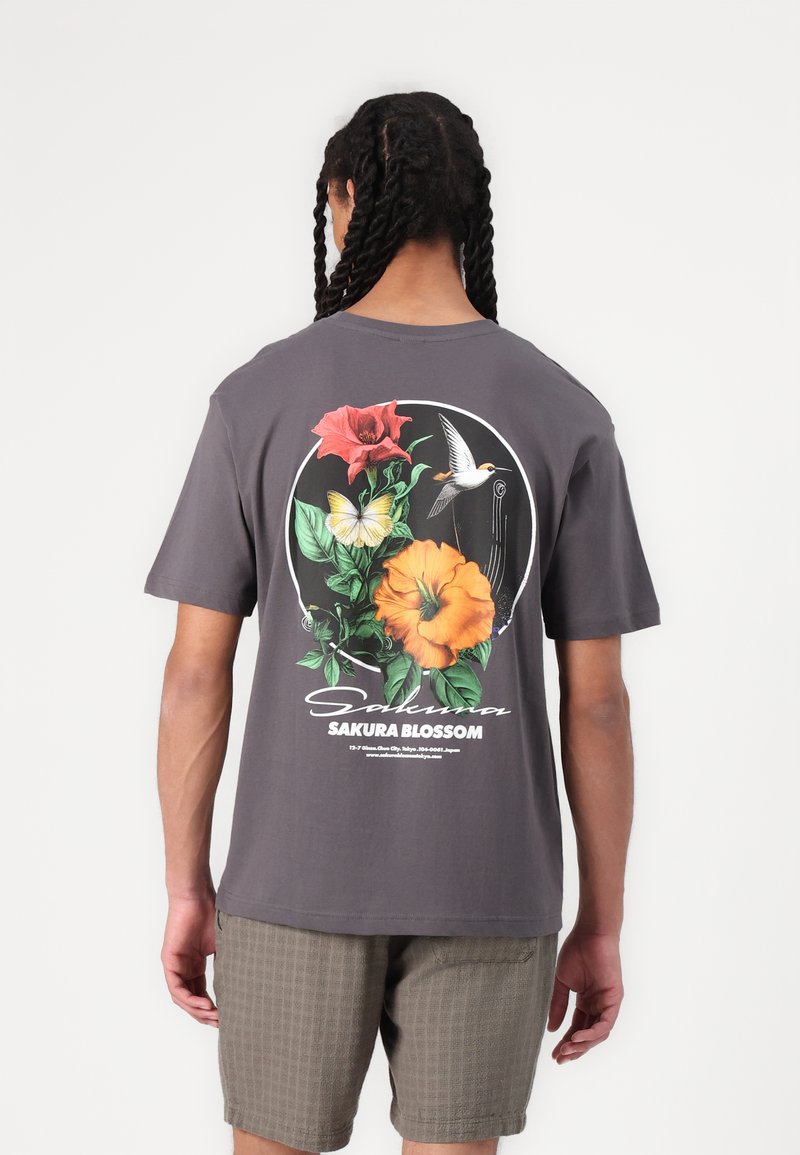 Mann trägt graues T-Shirt mit buntem Blumen-, Schmetterlings- und Vogeldesign sowie dem Text "Sakura Blossom" auf der Rückseite.