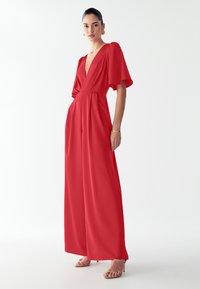 Kvinde iført en rød bredbenet jumpsuit med dyb V-udskæring og lette ærmer, parret med beige stroppede hæle og guld hoops øreringe.