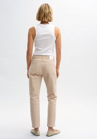 Top bianco a coste, pantaloni beige slim-fit con tasche posteriori, orli piegati e scarpe slip-on chiare su uno sfondo neutro.
