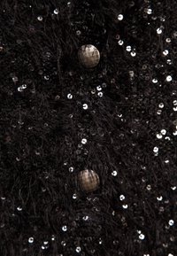 Svart texturerad tyg prytt med paljetter, försedd med två fasetterade metallknappar. Närbilden framhäver skinande och fnöskiga element.