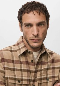 Camisa de franela a cuadros en beige y marrón con cierre de botones, dos bolsillos en el pecho y cuello clásico, usada sobre una prenda superior de color claro.