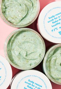 Groene hoofdhuid scrub met een romige textuur en spikkels. Verpakt in een doorzichtige container met een witte deksel, met blauwe tekst op het etiket.