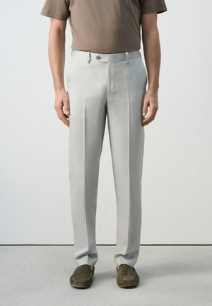 Hombre con pantalones formales gris claro, camisa de manga corta marrón y mocasines marrón oscuro, de pie frente a un fondo claro y liso.