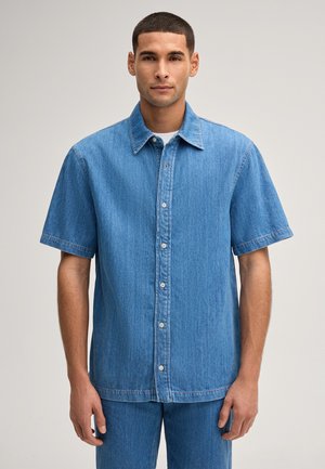 Mann steht und trägt ein blaues, kurzärmliges Jeanshemd, das zugeknöpft ist, mit passenden Jeanshosen vor einfachem Hintergrund.