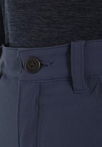 Nahaufnahme des Taillenbunds dunkler Hosen mit schwarzem Knopf und Gürtelschlaufe, kombiniert mit einem dunkel melierten Hemd, das eingesteckt ist.