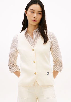 Frau mit langen dunklen Haaren, die eine cremefarbene, geknöpfte Weste über einem beige gestreiften Hemd und passenden cremefarbenen Hosen trägt, steht mit den Händen hinter dem Rücken.