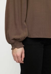 Brun transparent blus med voluminösa ärmar, featuring smockade manschetter med en ruffled kant. Matchad med svarta åtsittande byxor.