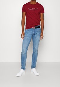 Rotes Baumwoll-T-Shirt mit "HILFIGER NEW YORK" in Weiß bedruckt. Blaue Jeans und weiße Sneakers vervollständigen das Outfit, das mit einem schwarzen Gürtel ausgestattet ist.