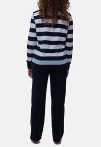 Osoba z kręconymi włosami stoi boso tyłem, ubrana w długą, niebiesko-białą koszulę w paski i ciemne spodnie na białym tle.