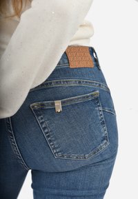 Jeans in denim blu con un design aderente, dotati di una toppa in pelle marrone con il logo in vita e di un accento in metallo argentato su una tasca posteriore.
