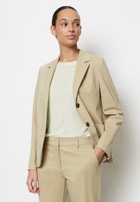Blazer beige con due bottoni e rever a notched, abbinato a una t-shirt verde chiaro. Vestibilità sartoriale, tessuto liscio. I pantaloni sono dello stesso colore del blazer.