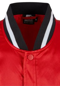 Rød bomberjakke med en glat tekstur, der har en sort krave med hvide striber og lukning foran med trykknapper.