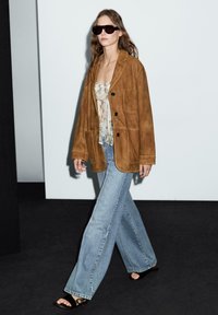 Blazer oversize in suede marrone, canotta fantasia, jeans a gamba larga azzurri e sandali neri con cinturini. Modella con capelli lunghi e occhiali da sole.