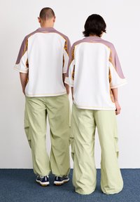 Twee individuen dragen losse, color-blocked jerseys, voornamelijk wit met lavendel en gele accenten, gecombineerd met lichtgroene cargobroeken.
