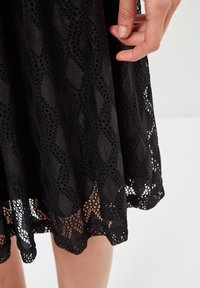 Vestido de encaje negro con una textura de patrón de diamantes, que presenta un dobladillo decorativo. Exhibe recortes intrincados y un acabado de tela suave.