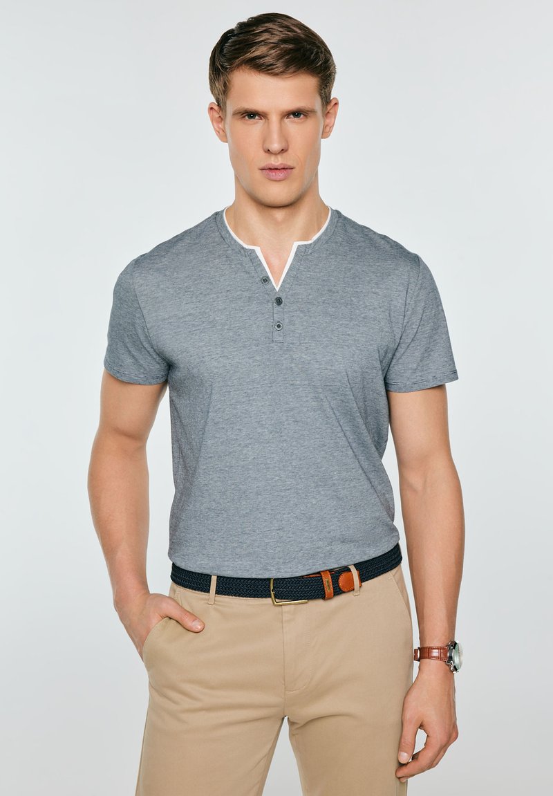 Lühikeste pruunide juustega mees, kes kannab halli lühikeste varrukatega henley-tüüpi särki, beežid pükse, musta vööd ja käekella ning seisab ühe käega püksitaskus.