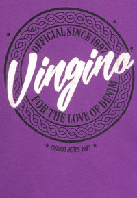 Fialové bavlněné tričko s kulatým potiskem, na kterém je v bílé tučné kurzívě napsáno "Vingino" s černým okrajem. Text obsahuje "Oficiálně od 1997."