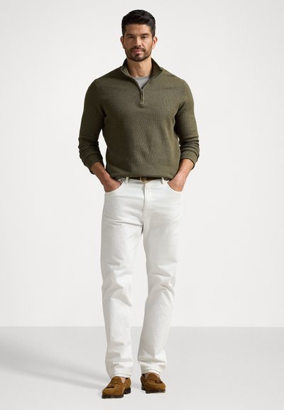 Sweater verde-oliva com meia-zíper, calças de ganga crèmes e mocassins castanhos. O suéter tem punhos e gola canelados; as calças são de perna reta e texturizadas.