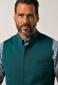 Turkos ärmlös väst med hög krage, med en knappfront, en sidoficka och en kontrasterande grön rutig skjorta under.