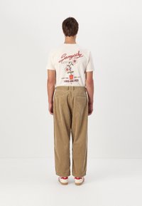 Pantalones de pana beige con un ajuste relajado, combinados con una camiseta crema que presenta gráficos de flores coloridas y texto. Zapatos con acentos rojos.