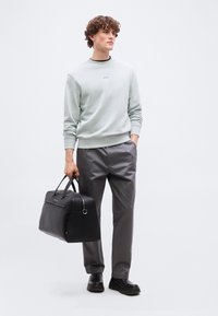 Νεαρός άνδρας με σγουρά μαλλιά, φοράει ανοιχτό γκρι φούτερ και σκούρο παντελόνι, κρατώντας μια μαύρη δερμάτινη τσάντα χειρός στο αριστερό του χέρι.