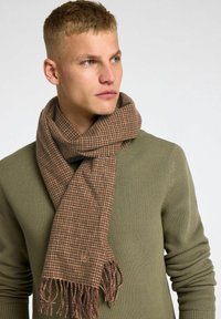 Scarf a quadri marrone realizzato in tessuto morbido, con orli sfrangiati, indossato in modo sciolto attorno al collo sopra un maglione verde.