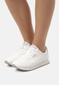 Zapatillas deportivas blancas de material sintético, con un upper texturizado, lengüeta acolchada y sutiles acentos grises en el talón y el logotipo.