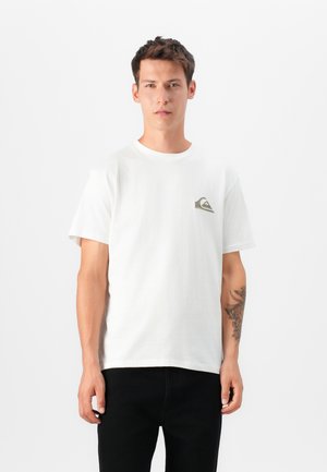 Nuori mies seisoo kasvo edellä, pukeutuneena valkoiseen t-paitaan, jossa on pieni logo rinnassa, ja mustiin housuihin, vasemmassa kyynärvarressa tatuointi.