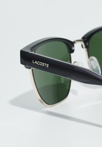 Ochelari de soare negri cu lentile verzi, având un accent auriu pe rame și un model texturat de culoare purpurie închis pe brațe. Brand: Lacoste.