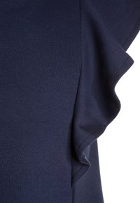 Granatowa bluzka z fakturowanej tkaniny, z falbanami na rękawach. Gładka powierzchnia z delikatnym prążkowanym wzorem na brzegach.