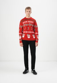 Jonge man staat in een rode feesttrui met snoepstokken, hulst, kerstman gezichten en de tekst "Blissful Holidays", zwarte broek en sneakers.