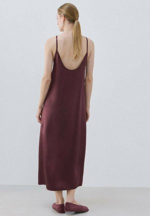 Vrouw met blond haar in een paardenstaart, gekleed in een mouwloze maxi-jurk in diep bordeauxrood en bijpassende slippers, staand tegen een effen grijze achtergrond.