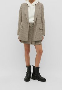 Veste à carreaux marron clair et noir portée sur un sweat à capuche blanc, associée à une mini-jupe assortie et des bottines noires à boucle.