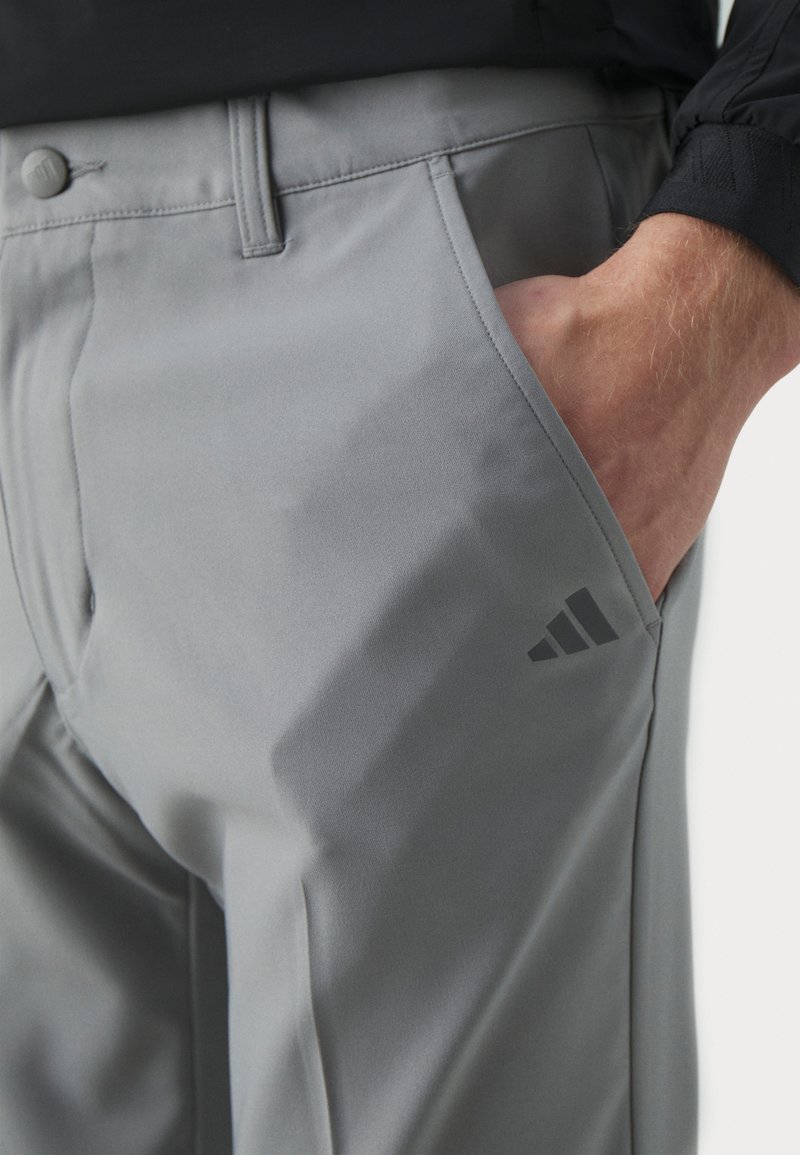 Graue Sporthose aus glattem Stoff, mit Knopfschluss, Seitentaschen und einem dezenten schwarzen Logo in der Nähe des Taillenbundes.