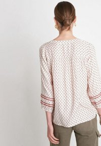 Blouse de couleur claire avec un motif floral rouge, dotée de manches trois-quarts, d'un col rond et d'ornements brodés sur les poignets.