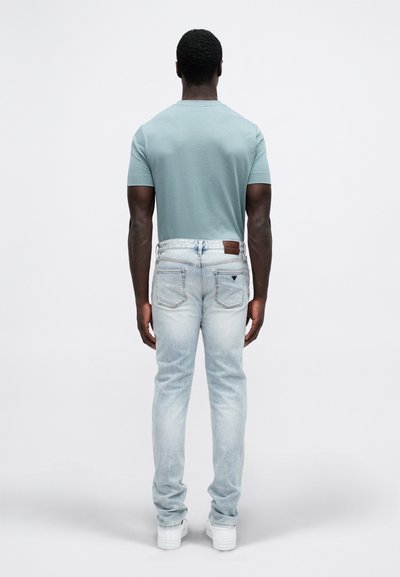 Mann med mørk hud iført lyseblå jeans, skjorte med korte ermer i lys tealfarge og hvite joggesko, som står vendt bort fra kameraet mot en ensfarget hvit bakgrunn.