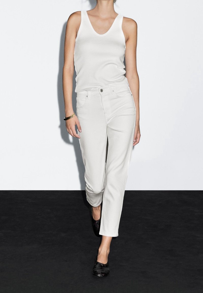 Femme portant un débardeur blanc et un jean blanc taille haute, chaussures plates noires et un bracelet en or, debout contre un mur blanc.