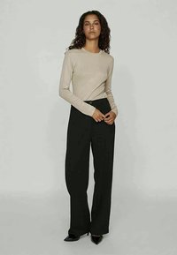 Woman stands wearing beige long-sleeve top, high-waisted black wide-leg pants, and black pointed shoes against plain gray background.